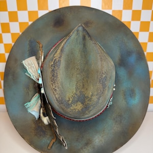 May include: A teal-coloured hat with a wide brim and a pinched crown. The hat features a red band and is adorned with feathers, turquoise beads, and fabric accents. The hat has a vintage, weathered appearance. The hat's brim measures approximately 30 cm in diameter.