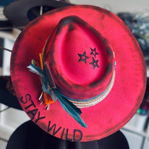 May include: A bright pink fedora hat with the words "STAY WILD" and three black star designs. The hat has a decorative band with feathers and trim.