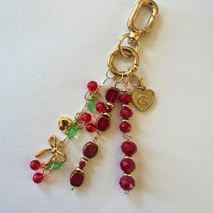 May include: A gold-toned keyring featuring a lobster clasp and a large ring. Dangling from the ring are three strands of red faceted beads, small green leaf-shaped beads, a gold-toned bow charm, a small gold heart, and a heart charm with a letter G.