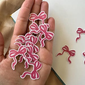 Puede incluir: Un conjunto de pegatinas de lazo rosa con contornos blancos. Los lazos, de varios tamaños, están en una mano. Las pegatinas tienen un acabado brillante, posiblemente de vinilo. Los lazos son de un tono rosa brillante, ideales para manualidades.