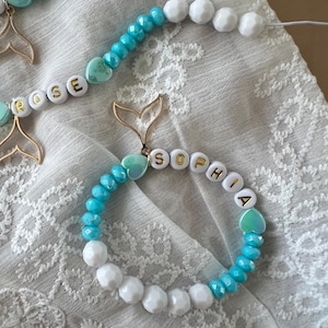 Könnte beinhalten: Ein Armband und ein Schnullerband mit weißen und türkisfarbenen Perlen. Das Armband schreibt "SOPHIA" mit goldenen Buchstabenperlen und hat einen goldfarbenen Meerjungfrauenschwanz-Anhänger und eine herzförmige Perle. Das Schnullerband schreibt "ROSE".