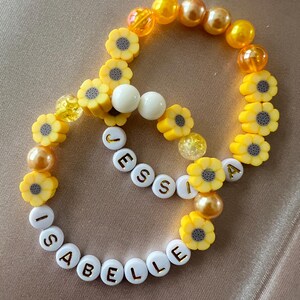 Könnte beinhalten: Zwei Armbänder mit Perlen. Ein Armband schreibt "Isabelle" und das andere "Jessia". Beide Armbänder haben gelbe Sonnenblumenperlen, weiße Perlen und gelbe und goldfarbene Perlen.