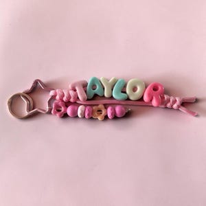 May include: A keyring featuring a pink star-shaped metal clasp attached to a pink cord. The cord holds pastel-coloured letter beads spelling TAYLOR in pink, blue, and cream, alongside a second strand of pink beads in various shapes and sizes.