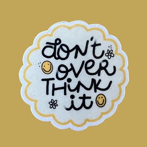 Puede incluir: Una pegatina blanca con borde festoneado con la frase "don't over think it" en letras negras. El diseño incluye dos caras sonrientes y dos pequeñas ilustraciones de flores. El fondo es de color amarillo mostaza liso.