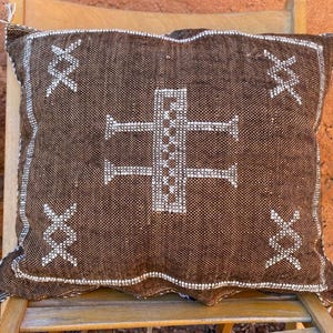 Peut inclure: Coussin décoratif marron avec des motifs géométriques blancs. Le coussin présente un motif central en forme de croix et des coutures décoratives sur les bords. Le coussin est posé sur une chaise en bois.