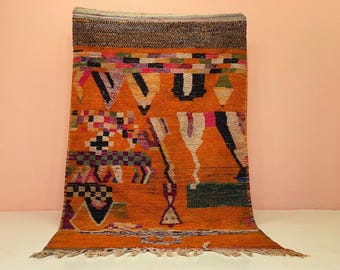 Puede incluir: Una vibrante alfombra naranja con patrones geométricos en negro, blanco, rosa, verde y morado. La alfombra tiene una superficie texturizada y un borde con flecos. El diseño incluye triángulos, cuadrados y otras formas.