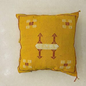 May include: Mustard yellow square cushion with geometric patterns. The cushion features a central design with two upward-pointing arrows and a horizontal band, along with square patterns in each corner. The edges are trimmed with red stitching.
