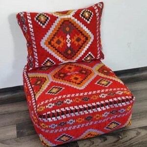 May include: A red upholstered chair with a matching pillow. The chair and pillow feature a geometric pattern in shades of orange, white, and black. The chair is square-shaped and appears to be made of a woven fabric.