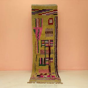 May include: A rectangular, olive green rug with a geometric design. The rug features pink, purple, and red shapes, with black and white accents. The rug has a fringe at the bottom and is displayed against a peach-colored wall.