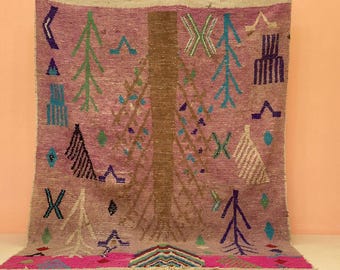 May include: A rectangular, hand-woven rug with a pink background and a central brown tree design. The rug features various geometric shapes and tree-like patterns in teal, green, white, and purple. The bottom edge has a bright pink border.