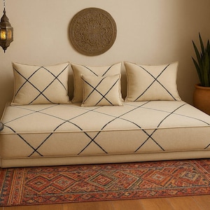 Puede incluir: Un sofá cama crema con costuras negras entrecruzadas, combinado con cojines a juego. Una alfombra estampada en tonos rojos, naranjas y marrones descansa sobre el suelo de madera. Una decoración de pared y una lámpara colgante completan la escena.