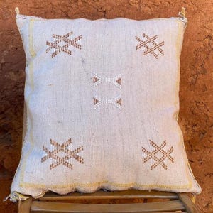 Può includere: Un cuscino decorativo quadrato bianco sporco con motivi geometrici marroni e bianchi. Il cuscino ha un bordo giallo e nappe agli angoli. È posto su una sedia di legno.