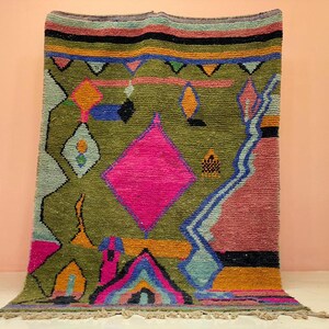 Peut inclure: Un tapis tissé à la main aux couleurs vives et aux motifs géométriques. Le tapis présente un fond vert olive avec un grand losange rose et des formes bleues, orange et noires. La bordure est ornée de rayures vertes, noires et oranges. Le tapis a une frange.