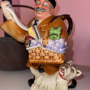 May include: A ceramic teapot figurine of a man wearing a brown coat and a black bowler hat, holding a newspaper and a basket with tea boxes. A white ceramic dog figurine is sitting at the man's feet.