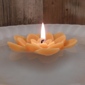 May include: A lit, orange lotus flower candle floats in a white bowl of water. The candle is shaped like a flower with multiple petals. The flame is bright and the candle is centered in the bowl.