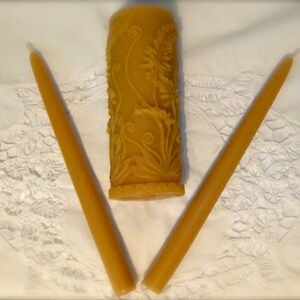 May include: Three beeswax candles on a white surface. Two long, slender tapers flank a cylindrical pillar candle. The pillar candle is decorated with a floral relief pattern. All candles are a warm, golden yellow color.