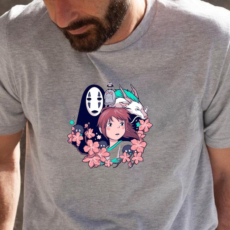 May include: A grey t-shirt with a vibrant anime-style graphic. The design features a character with brown hair, a dragon, and other figures surrounded by pink flowers. The t-shirt is made of a soft material.