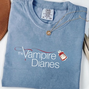 Puede incluir: Camiseta azul claro con el texto "The Vampire Diaries" en escritura blanca y roja. Un collar dorado con un peque&ntilde;o colgante descansa sobre la camiseta. La camiseta tiene una etiqueta Comfort Colors.