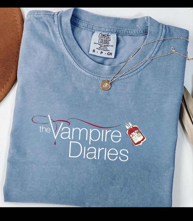 Puede incluir: Camiseta azul claro con el texto "The Vampire Diaries" en escritura blanca y roja. Un collar dorado con un peque&ntilde;o colgante descansa sobre la camiseta. La camiseta tiene una etiqueta Comfort Colors.