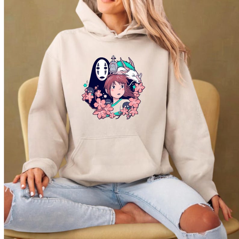 May include: Beige hoodie with a colourful graphic of anime characters, including a girl, a dragon, and a faceless spirit, surrounded by pink flowers. The hoodie has a front pocket and a drawstring hood.