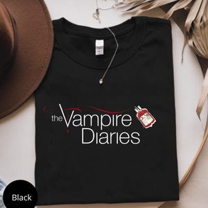 Puede incluir: Camiseta negra con el texto "the Vampire Diaries" y un gr&aacute;fico de bolsa de sangre. Un sombrero marr&oacute;n y un collar plateado son visibles en el fondo. La camiseta est&aacute; doblada.