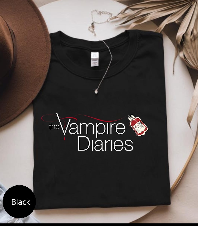 Puede incluir: Camiseta negra con el texto "the Vampire Diaries" y un gr&aacute;fico de bolsa de sangre. Un sombrero marr&oacute;n y un collar plateado son visibles en el fondo. La camiseta est&aacute; doblada.