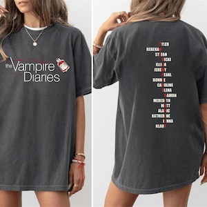 Puede incluir: Camiseta gris oscuro con el texto "the Vampire Diaries" en blanco y un gráfico rojo y blanco. La parte trasera de la camiseta muestra una lista de nombres en rojo y blanco.