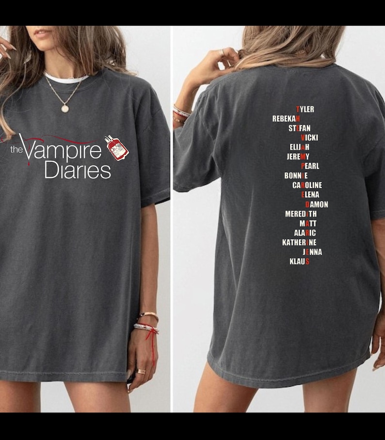 Puede incluir: Camiseta gris oscuro con el texto "the Vampire Diaries" en blanco y un gr&aacute;fico rojo y blanco. La parte trasera de la camiseta muestra una lista de nombres en rojo y blanco.