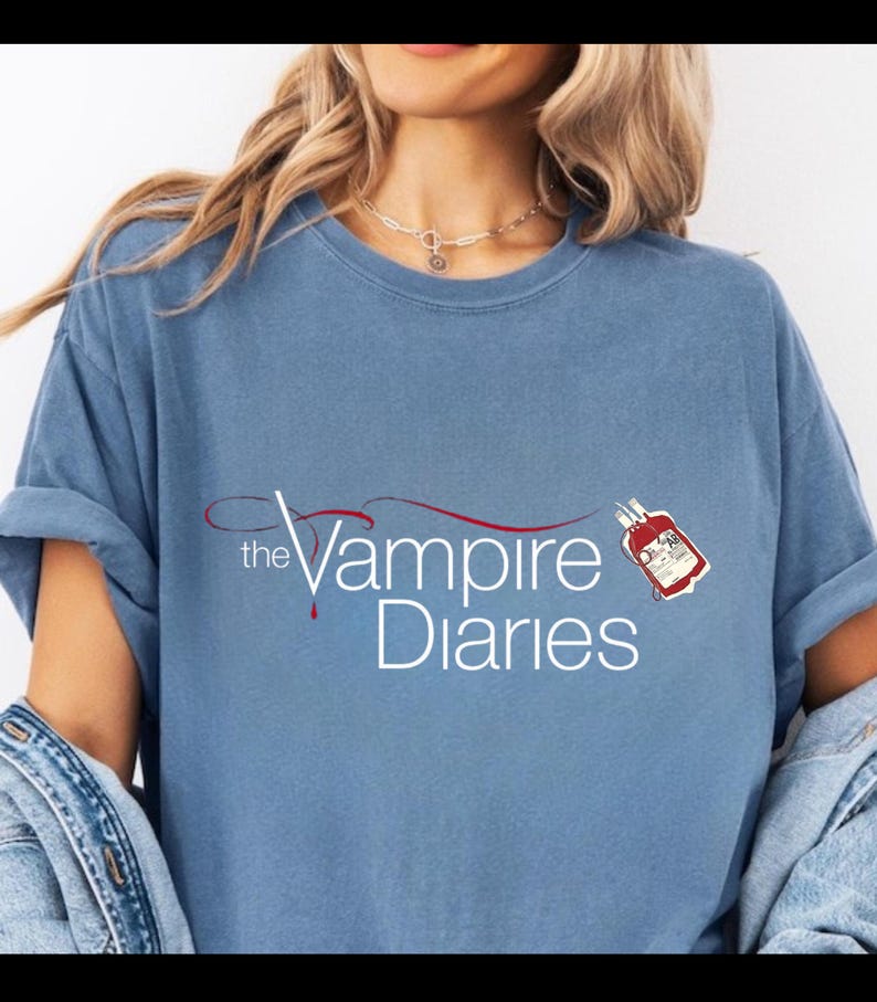 Puede incluir: Camiseta azul claro con la frase "the Vampire Diaries" en blanco y un dise&ntilde;o de bolsa de sangre roja. La camiseta tiene mangas cortas y un corte holgado, ideal para el uso diario.