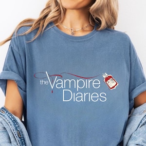 Puede incluir: Camiseta azul claro con la inscripción blanca "the Vampire Diaries" y un diseño de gota de sangre roja. La camiseta tiene mangas cortas y un corte holgado.