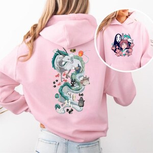 May include: Pale pink hoodie with a vibrant illustration of a dragon and other characters from an animated film. The back of the hoodie features a large dragon design, and the front pocket area has a smaller graphic of characters and flowers.