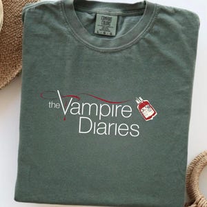 Puede incluir: Camiseta verde salvia con el texto blanco "the Vampire Diaries" y un gr&aacute;fico de bolsa de sangre roja. La camiseta est&aacute; doblada y tiene una etiqueta Comfort Colors.