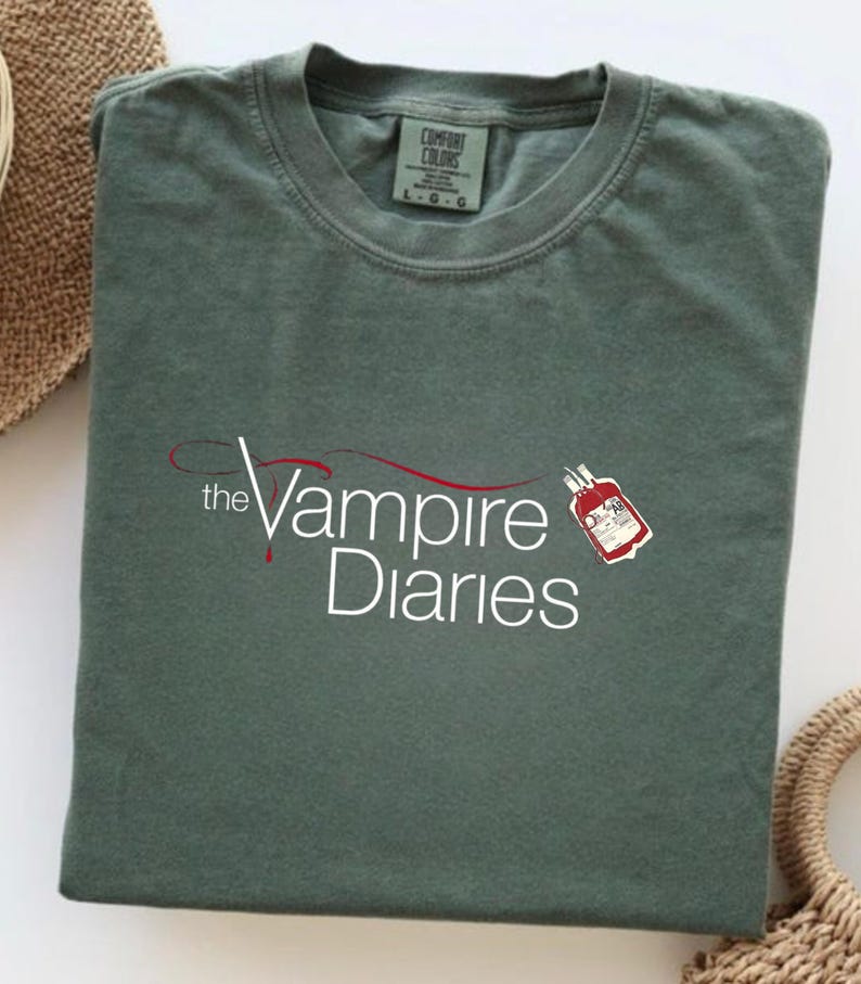 Puede incluir: Camiseta verde salvia con el texto blanco "the Vampire Diaries" y un gr&aacute;fico de bolsa de sangre roja. La camiseta est&aacute; doblada y tiene una etiqueta Comfort Colors.