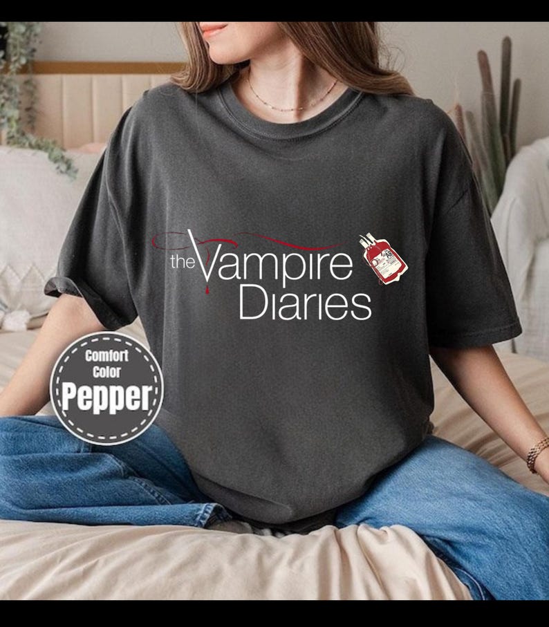 Puede incluir: Camiseta gris oscuro con el texto "the Vampire Diaries" en blanco y rojo. Un peque&ntilde;o gr&aacute;fico de una bolsa de sangre est&aacute; a la derecha. La camiseta est&aacute; etiquetada como "Comfort Color Pepper" en un dise&ntilde;o circular.
