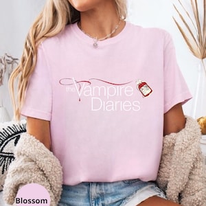 Puede incluir: Camiseta rosa claro con el texto "The Vampire Diaries" en una fuente estilizada, acompa&ntilde;ada de un dise&ntilde;o de gota de sangre roja. La camiseta es de estilo casual.
