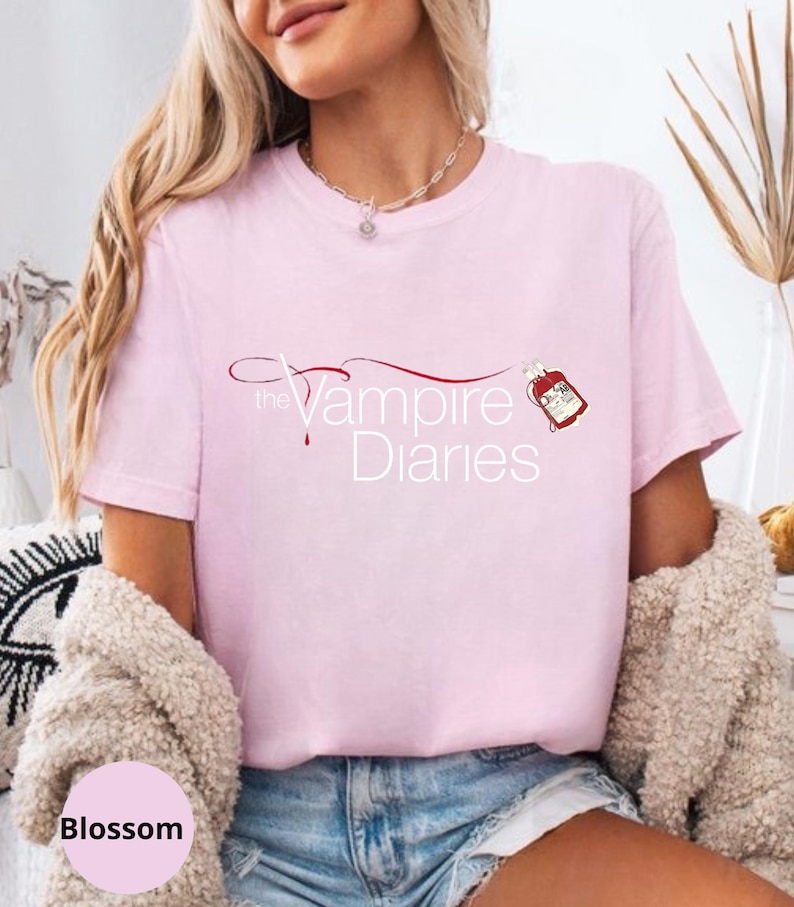Puede incluir: Camiseta rosa claro con el texto "The Vampire Diaries" en una fuente estilizada, acompa&ntilde;ada de un dise&ntilde;o de gota de sangre roja. La camiseta es de estilo casual.