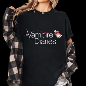Puede incluir: Camiseta negra con el texto "the Vampire Diaries" en escritura blanca y roja. Un peque&ntilde;o gr&aacute;fico de una bolsa de sangre es visible. La persona lleva una camisa a cuadros en blanco y negro encima.