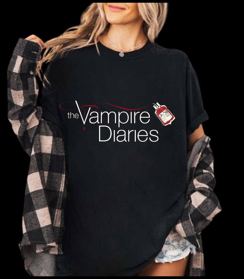 Puede incluir: Camiseta negra con el texto "the Vampire Diaries" en escritura blanca y roja. Un peque&ntilde;o gr&aacute;fico de una bolsa de sangre es visible. La persona lleva una camisa a cuadros en blanco y negro encima.