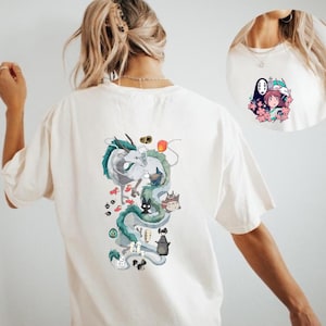 Op de afbeelding: Crèmekleurig T-shirt met een kleurrijke anime-stijl afbeelding op de rug. Het ontwerp bevat een draak, verschillende personages en andere elementen. Een kleinere afbeelding staat op de voorkant, met personages en bloemen.
