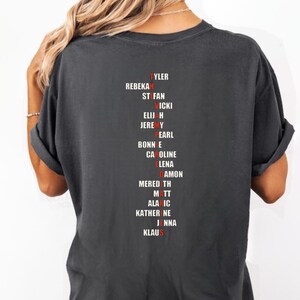 Puede incluir: Camiseta gris oscuro con una lista de nombres en fuente roja y blanca. Los nombres incluyen Tyler, Rebekah, Stefan, Vicki, Elijah, Jeremy, Pearl, Bonnie, Caroline, Elena, Damon, Meredith, Matt, Alaric, Katherine, Jenna y Klaus.