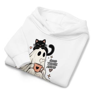 Può includere: Una felpa con cappuccio bianca con una stampa grafica raffigurante un fantasma che tiene in mano una tazza color pesca con un cuore, un gatto nero seduto sulla testa del fantasma e il testo "GOOD THINGS AHEAD" con piccoli cuori e stelle.