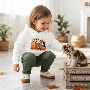 May include: A white hooded sweatshirt featuring a graphic print of three kittens, pumpkins, and autumn leaves with the text "Hello Fall". The sweatshirt is paired with olive green cargo pants and white sneakers, worn by a child.