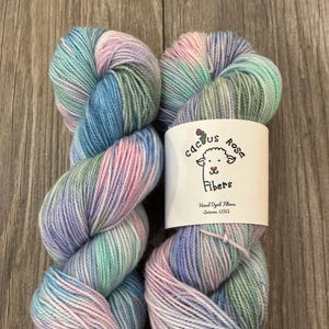 Puede incluir: Dos ovillos de hilo teñido a mano en una gama de colores con tonos de azul, verde, rosa y blanco. El hilo está etiquetado con "Cactus Rose Fibers, Hand Dyed Fibers, Arizona, USA."
