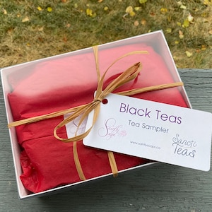 May include: A white gift box with red tissue paper inside, tied with a brown ribbon and a tag that reads "Black Teas Tea Sampler" with the website www.saintlysoaps.ca and the text "Saintly Teas".