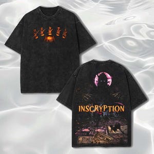 以下が含まれることがあります： グラフィックデザインの黒いTシャツ。前面にはオレンジ色の人物と中央のデザインが描かれています。背面にはオレンジ色の「INSCRIPTION」の文字と、生き物とカードゲームの画像が表示されています。このシャツはヴィンテージ感のあるウォッシュ加工が施されています。