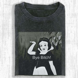 Op de afbeelding: Donkergrijs t-shirt met een zwart-witte afbeelding van een tekenfilmvrouw die zwaait, met de tekst "Bye Bitch!". De afbeelding is een parodie op een klassiek tekenfilmfiguur. Het shirt heeft een ronde hals en korte mouwen.