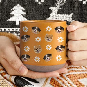 Könnte beinhalten: Beige Keramikbecher mit braunem Boden, verziert mit Bildern von Hunden und weißen Gänseblümchen. Der Becher wird von einer Person gehalten, die einen schwarz-weiß gemusterten Pullover trägt. Geeignet für Heißgetränke.