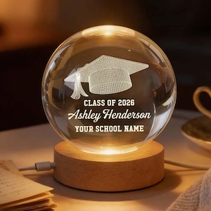 May include: A clear glass sphere with a graduation cap design, illuminated by a warm light. The sphere sits on a wooden base and features the text "CLASS OF 2026", a name, and "YOUR SCHOOL NAME".