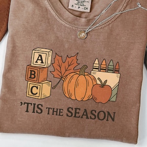 May include: Espresso-colored t-shirt with a fall-themed graphic. The design includes ABC blocks, a maple leaf, a pumpkin, an apple, and a box of crayons. The text "'TIS THE SEASON" is printed below the graphic. A silver necklace is draped across the shirt.