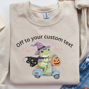 Puede incluir: Sudadera de color arena con un diseño temático de Halloween. El diseño presenta una rana verde con un sombrero de bruja y capa, montada en un scooter azul claro y sosteniendo una linterna de calabaza. El texto encima de la rana dice "Off to your custom text".